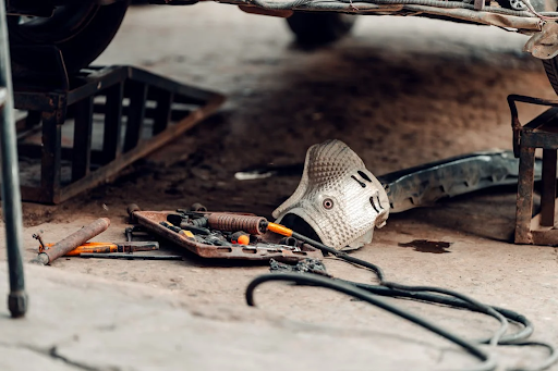 A workspace under a vehicle lifted on ramps, with scattered tools, a heat shield, and cables on the ground, indicating automotive repair or maintenance in progress.