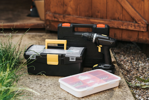 A set of tools placed outside a wooden shed, including a black and yellow toolbox, a power drill, and a plastic organizer containing screws and nails, ready for DIY or repair work