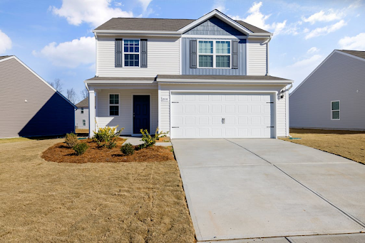 A two-story suburban house with white siding, blue shutters, and a large driveway leading to a double garage, surrounded by a neatly maintained lawn under a clear blue sky.