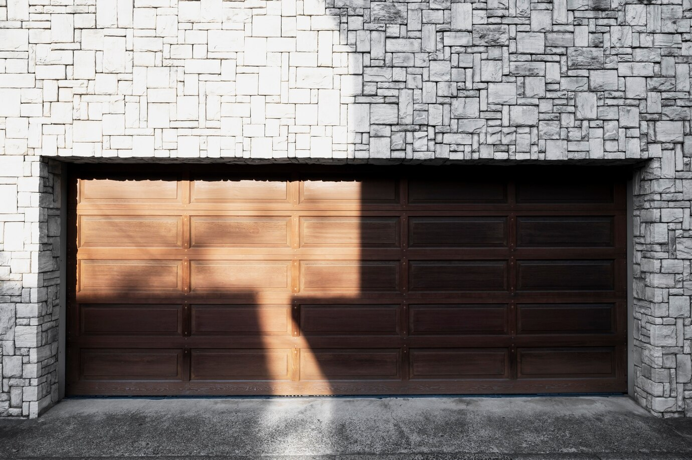 A wooden garage door with a paneled design set within a textured stone exterior wall, partially illuminated by sunlight casting sharp shadows