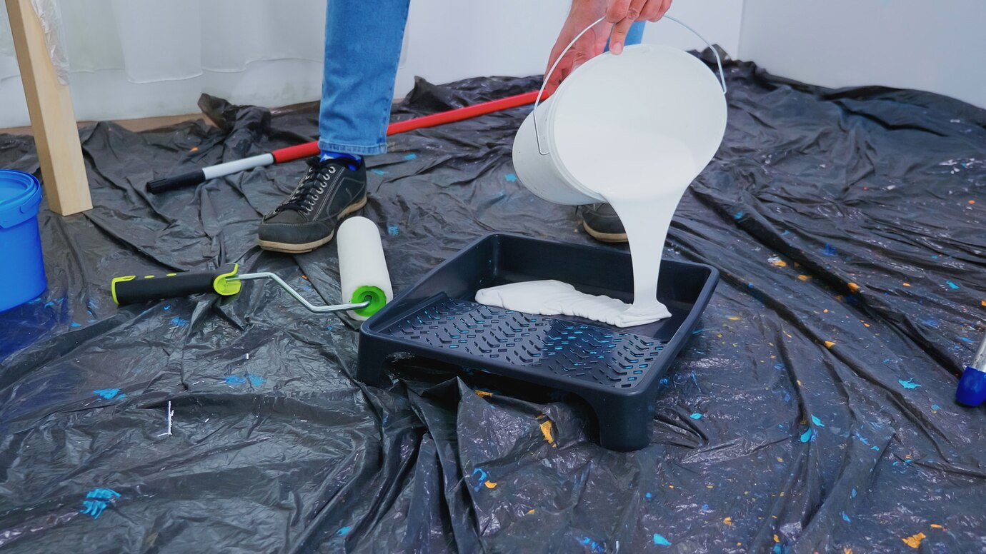 A person pouring white paint into a roller tray on a black protective tarp, with painting tools such as a roller brush and a paint bucket nearby, indicating a garage door renovation