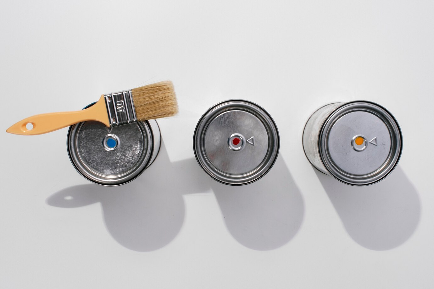 Three metal paint cans with color-coded labels on their lids, arranged in a row on a white surface. A paintbrush with a beige handle rests on the first can, casting a shadow on the surface