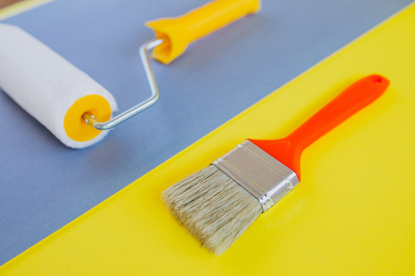 A close-up of a paint roller with a yellow handle and a brush with an orange handle, placed on a colorful surface divided into yellow and blue sections for garage door painting