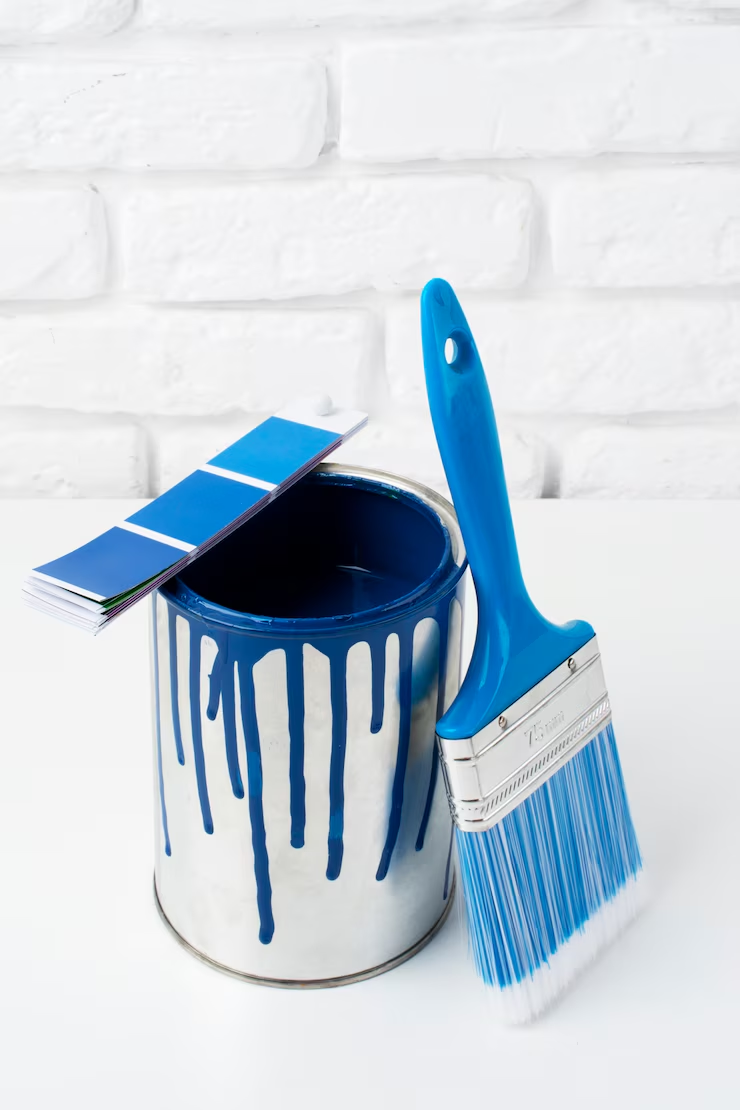 A metal paint can with blue paint dripping down the sides, accompanied by a blue paintbrush and a color swatch, set against a white brick background