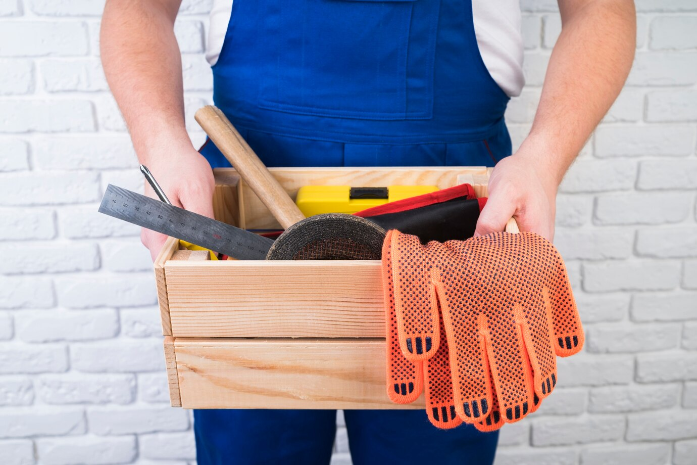 a person dressed in blue work overalls holding a wooden toolbox. The toolbox contains various tools such as a ruler, a hammer, and other work accessories. Bright orange safety gloves with black dotted grip are draped over the edge of the toolbox