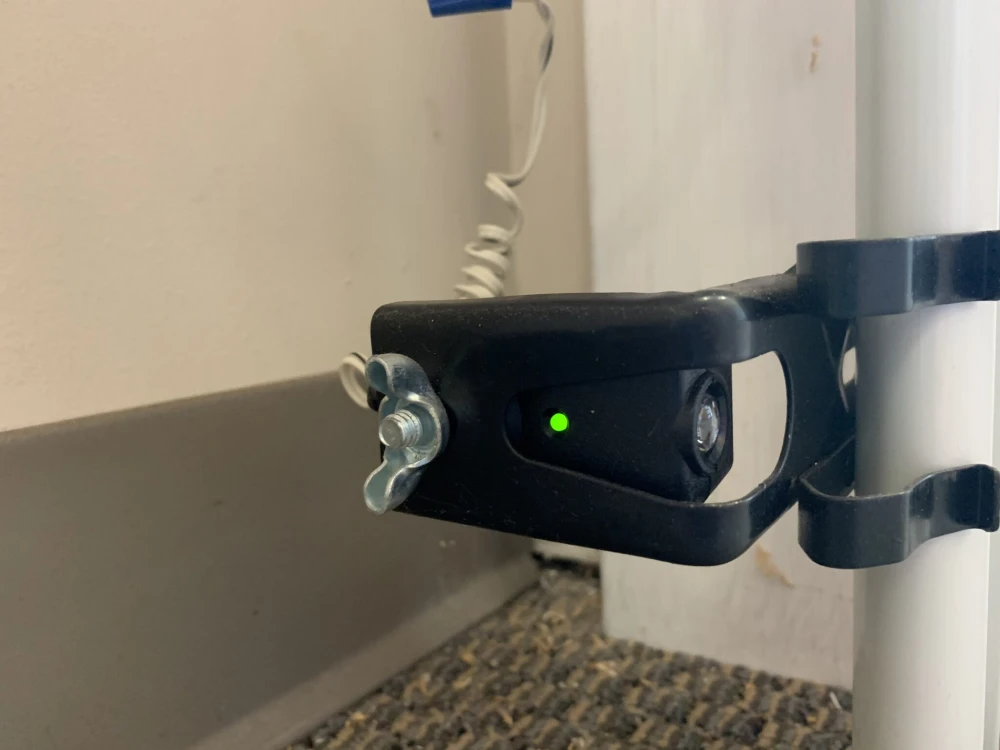 A close-up image of a black garage door safety sensor mounted on a white metal bracket with a green indicator light turned on, indicating proper alignment