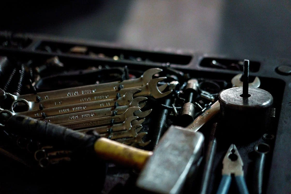 a collection of tools, including wrenches, bolts, and other mechanical items, likely arranged in a toolbox or workbench tray. The setting appears dimly lit, emphasizing the metallic sheen of the tools. The organized placement of the wrenches suggests readiness for mechanical or repair work