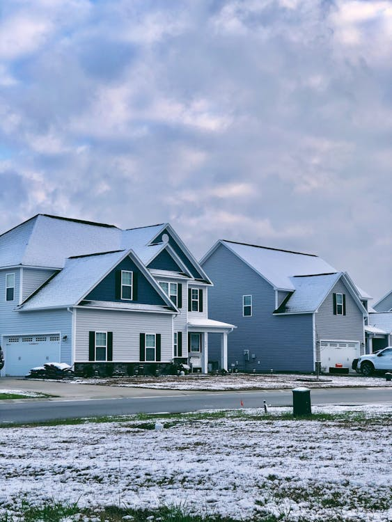 a suburban neighborhood in winter, featuring two modern-style houses with light-colored siding and steeply sloped roofs partially covered in snow. The foreground highlights a lightly snow-dusted lawn and a paved driveway, while the sky above is cloudy with patches of blue peeking through. A white car is parked near one of the homes, emphasizing the peaceful residential setting. The overall atmosphere suggests a calm, cold day in a well-maintained community