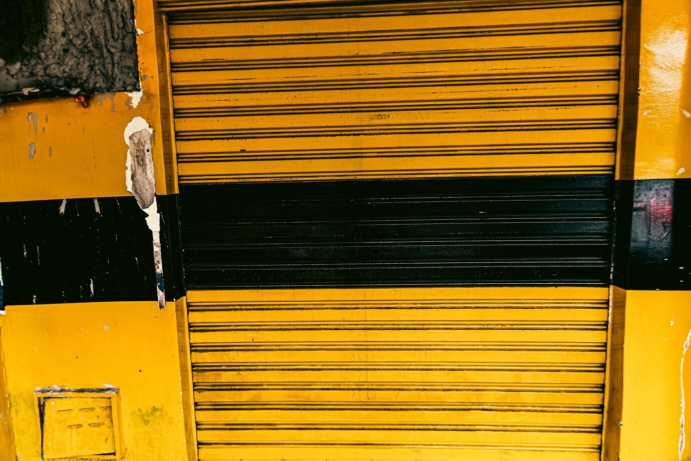 Сlose-up of a yellow metal roller shutter door with a bold black stripe running horizontally through the center. The shutter shows signs of wear, with chipped paint and exposed material in some areas, giving it a rugged and industrial aesthetic. The textures and colors create a striking visual contrast