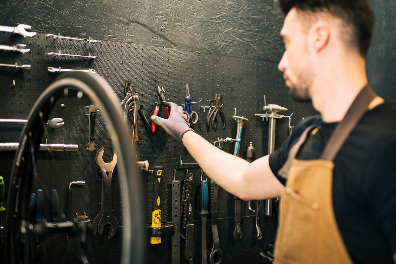 A workshop setting where a person, wearing a brown apron and gloves, is reaching for a tool from a pegboard mounted on a dark wall. The pegboard is organized with various tools such as pliers, wrenches, and screwdrivers. A bicycle wheel is partially visible in the foreground, suggesting a bicycle repair or maintenance context. The lighting creates a focused, professional atmosphere, highlighting the importance of tools in the workspace