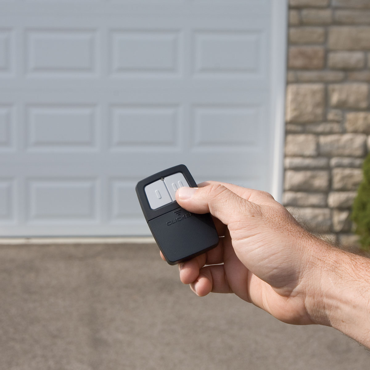 A hand holding a black garage door remote control in front of a closed white garage door with a stone wall in the background