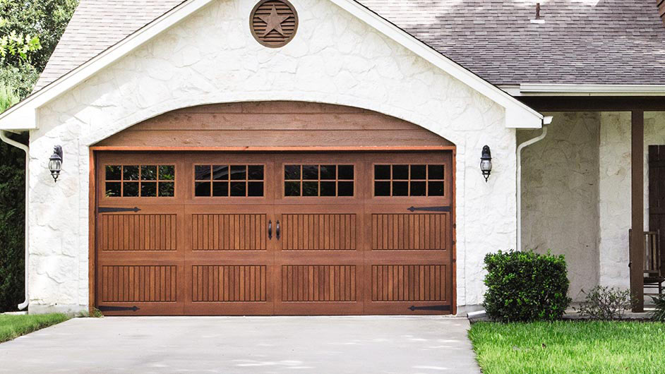 a beautifully designed carriage-style garage door with a wood-look finish. The door is divided into symmetrical panels with vertical slats and decorative hardware, giving it a traditional and elegant appearance