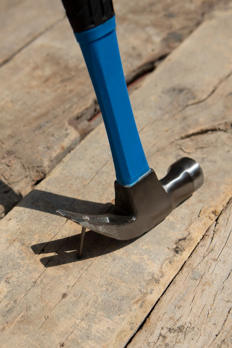 А hammer with a blue handle being used to remove a nail from a wooden surface. The hammer's claw is engaged with the nail, and the wood shows signs of wear and natural texture, suggesting a rustic or outdoor setting. The lighting emphasizes the tool's metallic surface and the grain of the wood