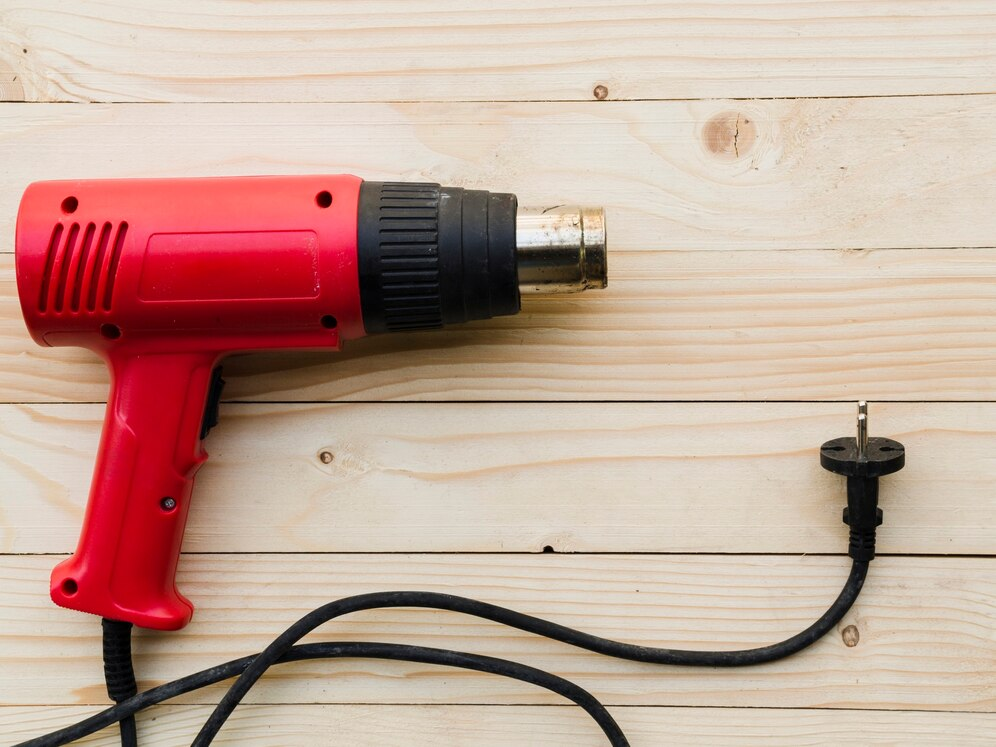 А red heat gun placed on a wooden surface. The heat gun has a metallic nozzle and a black handle grip, with its cord and plug visible in the foreground. The light-colored wooden background highlights the vibrant red color of the tool and its clean, simple design. The image emphasizes the functionality of the heat gun, often used for DIY projects or repairs