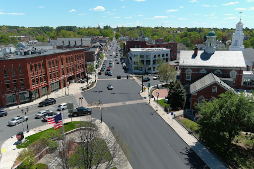 downtown area of Andover, MA, from an elevated perspective. The scene features a prominent intersection surrounded by a mix of historical red-brick buildings, modern structures, and classic New England architecture, including a church with a white steeple on the right