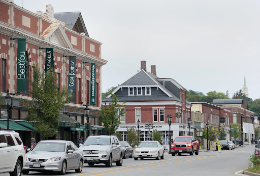 a lively street scene in Andover, MA. The view features a charming downtown area with a mix of historical brick buildings and modern storefronts. Prominent businesses, such as cafes and retail stores, line the street, with signs and banners adding color and character