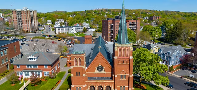 An aerial view of Arlington local area featuring a prominent red-brick church with a tall green steeple in the center. The surrounding neighborhood includes residential homes, apartment buildings, and landscaped greenery under a clear blue sky, showcasing a blend of urban and suburban charm