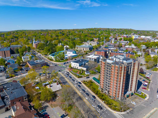 An aerial view of Arlington local area showcasing a mix of residential and commercial buildings surrounded by lush greenery. The scene includes a tall apartment building in the foreground, busy streets with traffic, and several notable landmarks, such as a church and other historical buildings, under a bright blue sky