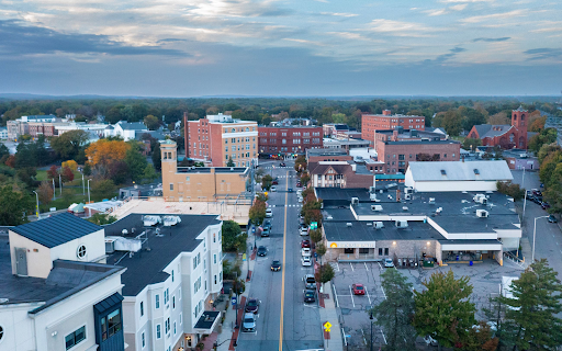 An aerial view of downtown Attleboro, MA, showcasing a mix of modern and historic brick buildings under a moody, cloudy sky. The central street is lined with trees in autumn colors and cars parked along both sides. The scene highlights the blend of commercial and residential spaces with a small-town charm