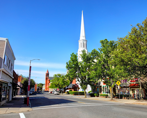 A vibrant street view in Beverly, MA, featuring the iconic white spire of a historic church surrounded by lush green trees. The street is lined with quaint shops and local businesses, leading to a red brick church in the distance. A clear blue sky provides a bright and cheerful backdrop, highlighting the charm of this small New England town