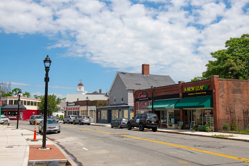 A picturesque street view in Beverly, MA, showcasing a blend of local businesses, including 'A New Leaf,' housed in charming, historic-style buildings. The scene features a clean sidewalk, classic lampposts, and parked cars, all set against a backdrop of lush green trees and a bright blue sky with scattered clouds. The white dome of a historic building peeks out in the distance, adding to the small-town charm