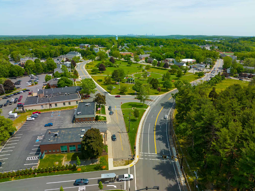 Aerial view of Burlington featuring a central green park surrounded by winding roads and buildings. The area includes trees, parking lots, and a mix of commercial and residential structures. A clear, sunny sky adds brightness to the scene