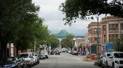 a street-level view of Chelsea, MA. The scene shows a bustling urban street with parked cars lining both sides of the road. In the background, the iconic Tobin Bridge dominates the scene, visible through the line of buildings and trees