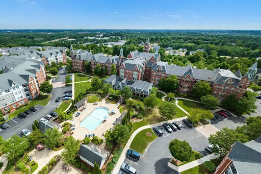 Aerial view of a residential complex in Danvers, MA, featuring red brick buildings with gabled roofs. The area includes a central swimming pool, green landscaped gardens, and a parking lot. Surrounding trees and a distant horizon add to the scenic view.