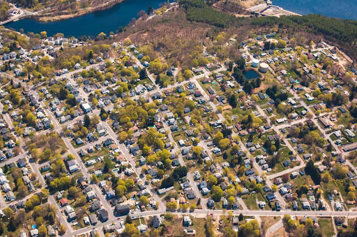 An aerial view of Danvers, MA, showing a suburban neighborhood with rows of houses surrounded by trees. A river and a forested area are visible in the background, along with a water tower.