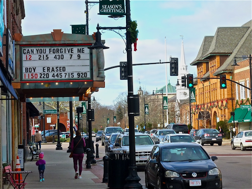 view of Dedham, MA, captures a lively downtown scene with pedestrians, cars, and a theater marquee displaying movie titles