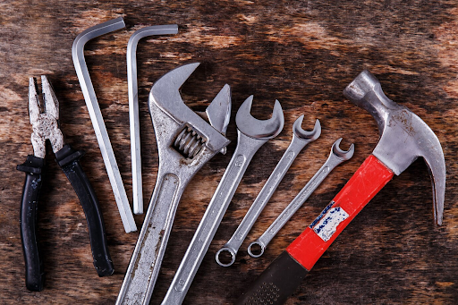 An assortment of hand tools including pliers, Allen wrenches, an adjustable wrench, a set of combination wrenches, and a red-handled claw hammer arranged on a rustic wooden surface, showcasing signs of wear and frequent use