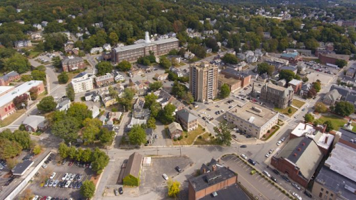 Aerial view of Fitchburg, MA, showcasing a mix of urban and residential areas. In the center, a tall building stands surrounded by smaller structures, including homes and commercial buildings. To the left, an industrial building and parking lot are visible, contrasting with the green, tree-covered areas in the background. The layout highlights the blend of historical architecture and modern infrastructure characteristic of the city