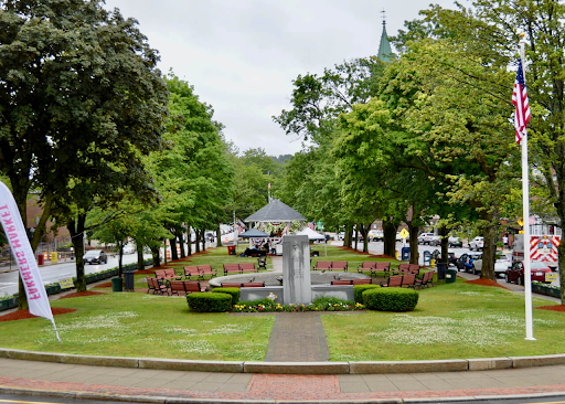 Central park area in Fitchburg, MA, featuring a well-maintained green lawn surrounded by trees and red-brick pathways. A central stone monument and circular fountain add a focal point to the park, with benches placed around for visitors to relax. An American flag stands tall on the right, accompanied by a 'Farmer's Market' banner on the left. The backdrop includes a visible street with cars and a distant view of a church steeple, encapsulating the community-oriented charm of the area