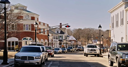 a street-level view of Lexington, MA, showcasing a bustling town center. The scene highlights a blend of historical and modern architecture, with a prominent red-brick building featuring a clock tower on the left. Traditional white clapboard buildings line the street, reinforcing the New England charm of the area