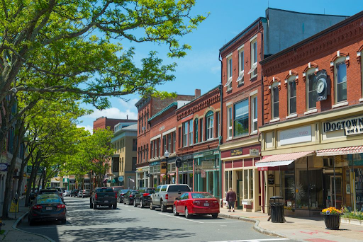 A vibrant downtown street in Gloucester, MA, with historic brick buildings, shops, and cars parked along the sides