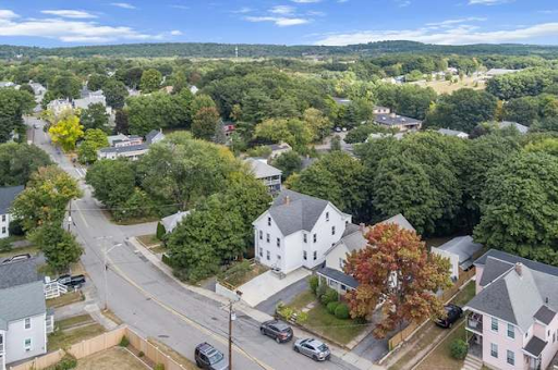 A scenic aerial view of a residential neighborhood in Leominster, MA, featuring charming homes with gabled roofs surrounded by lush greenery and mature trees. The quiet streets and well-maintained yards create a serene suburban atmosphere, while the rolling hills and expansive green spaces in the background highlight the area's natural beauty under a bright, partly cloudy sky