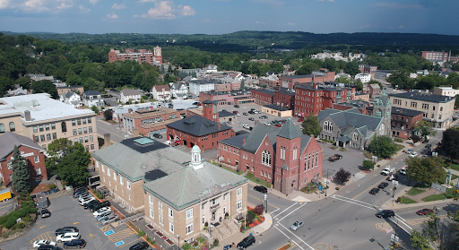 Aerial view of Leominster, MA, highlighting its historic downtown area with prominent brick buildings, a traditional town hall with a white cupola, and churches with distinctive architecture. The photo showcases a mix of residential and commercial structures surrounded by greenery, with rolling hills in the background under a partly cloudy blue sky, reflecting the town's blend of urban and rural charm