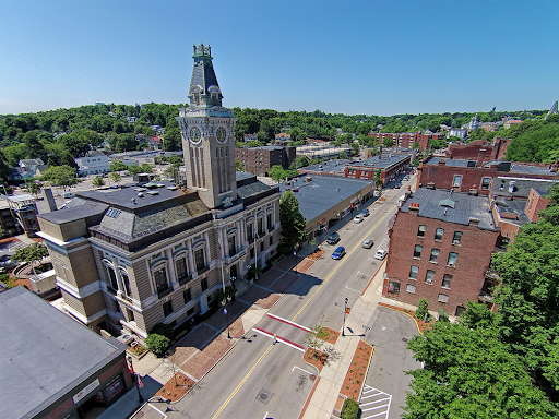 Aerial view of downtown Marlborough, MA, featuring a historic building with a tall clock tower along a main street lined with brick structures. The area is surrounded by lush green hills under a clear blue sky, highlighting the town’s blend of architectural heritage and scenic charm