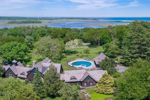 an aerial view of Marshfield, MA, featuring a sprawling estate surrounded by lush greenery. The scene includes a circular swimming pool, expansive lawns, and a backdrop of coastal wetlands and ocean views