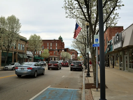 Street view of Milford, MA, showcasing a busy main street lined with parked cars, brick buildings, American flags, and blooming trees on both sides