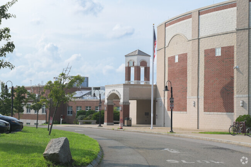 A brick and cream-colored building in Milton, MA, featuring a clock tower with arches. The area includes a circular driveway, a grassy lawn with trees, and an American flag pole in front of the building on a partly cloudy day