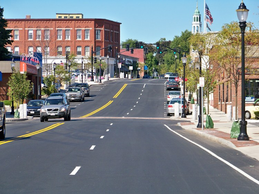 Reading, MA, featuring a wide street lined with small businesses, trees, and parked cars. A red brick building stands prominently on the left, while a church with a tall steeple and American flag are visible in the background