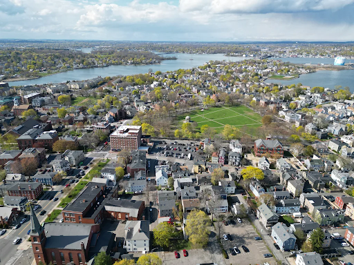 Aerial view of Salem, MA, showcasing a blend of historic and residential architecture surrounded by lush greenery. In the foreground, charming brick buildings and tree-lined streets are visible, while further back, a large green park and peaceful waterways frame the town. The photo captures the vibrant, small-town charm of Salem with a backdrop of expansive blue skies and scattered clouds