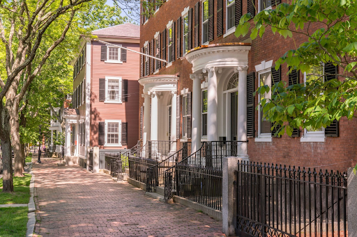 Charming historic street in Salem, MA, featuring brick sidewalks and classic New England-style homes with ornate white columns and black shutters. The street is shaded by lush green trees, adding a sense of tranquility and charm. The sunlight filters through the leaves, casting dappled shadows on the sidewalk and enhancing the picturesque appeal of the scene