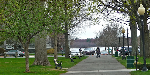 A park walkway in Wakefield, MA, lined with trees and benches on a cloudy day. People are walking and a stroller is visible on the pathway, with parked cars and a body of water in the background