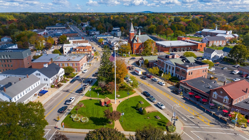 a vibrant aerial view of downtown Walpole, MA. At the center is a green park featuring a flagpole and landscaping, surrounded by roads, local businesses, and public buildings