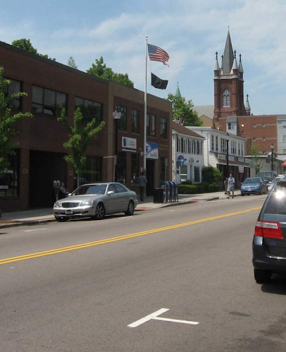 a street in Watertown Town, MA, featuring a blend of municipal and commercial architecture. Prominently visible is an American flag flying in front of a post office or civic building, indicating a community-centered area. The street is lined with small businesses housed in classic brick and white-painted buildings