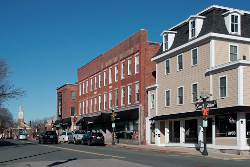 Street view of downtown Woburn, MA, featuring a row of historic red-brick buildings and modern beige structures under a bright blue sky. The buildings house local businesses, shops, and restaurants, with parked cars lining the street. Seasonal decorations adorn the lamp posts, adding charm to the urban scene. A church steeple is visible in the distance, adding to the town's quaint New England character