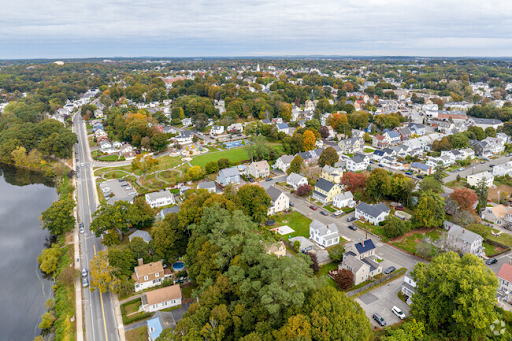 An aerial view of Woburn, MA, showcasing a charming suburban neighborhood with tree-lined streets and neatly arranged houses. A winding main road runs alongside a calm body of water on the left, leading to a community park with open green spaces and recreational areas. The landscape is dotted with a mix of autumnal and evergreen trees, reflecting the seasonal charm of the area under an overcast sky